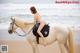 A woman in a bikini riding a white horse on the beach.